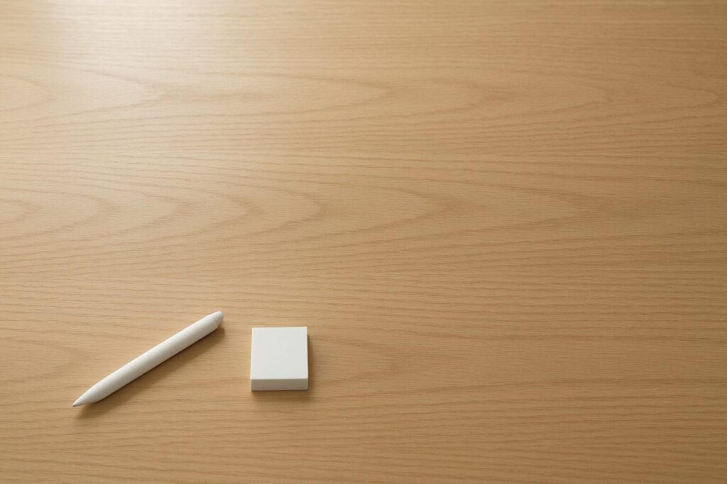 木目デスクを俯瞰した静かな作業シーン。手前に文具がそろい、奥に余白がある様子。